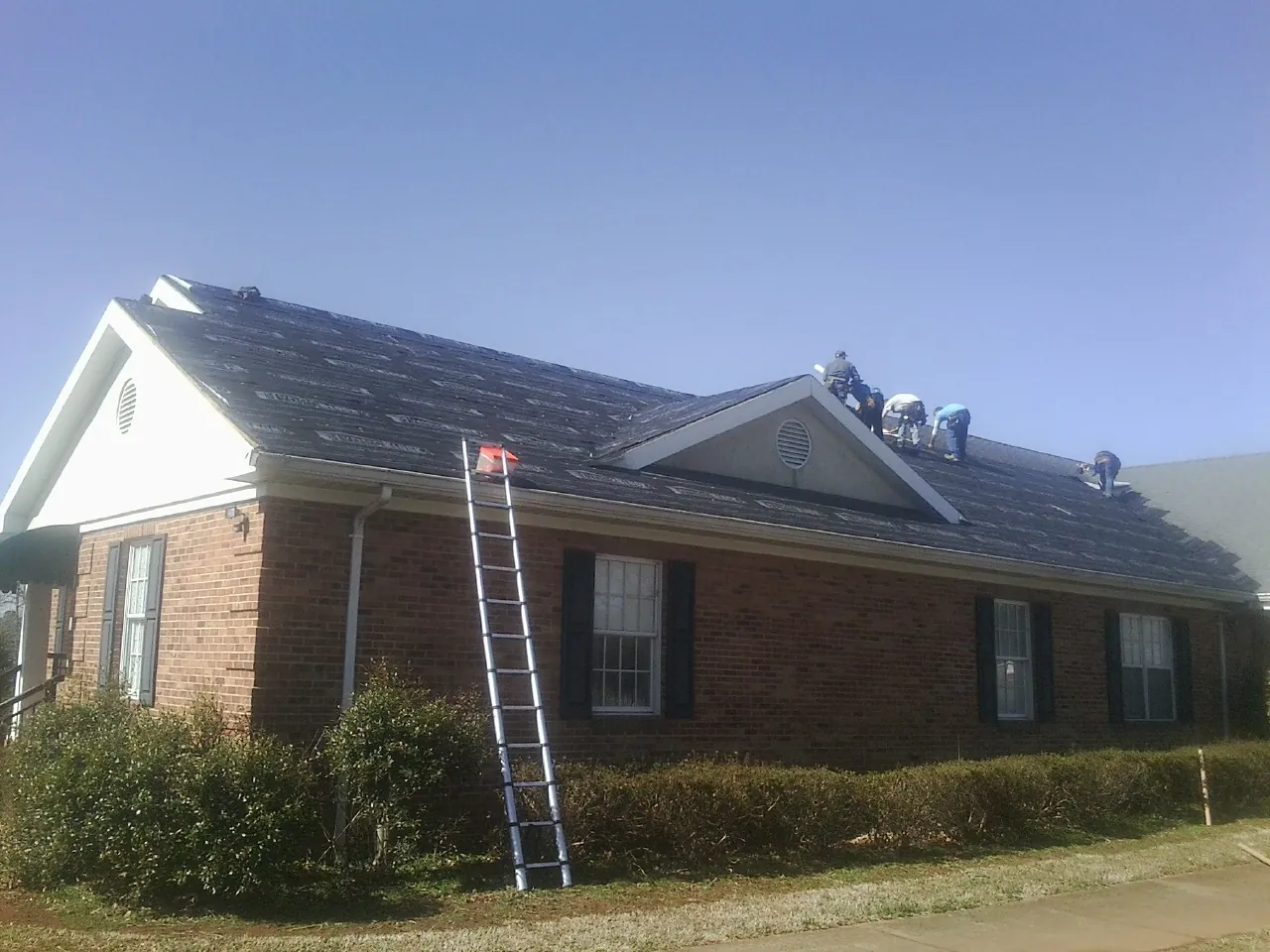 Crew tearing shingles off a brick ranch roof with a ladder set — Emergency Roof Repair work by our crew in Cologne