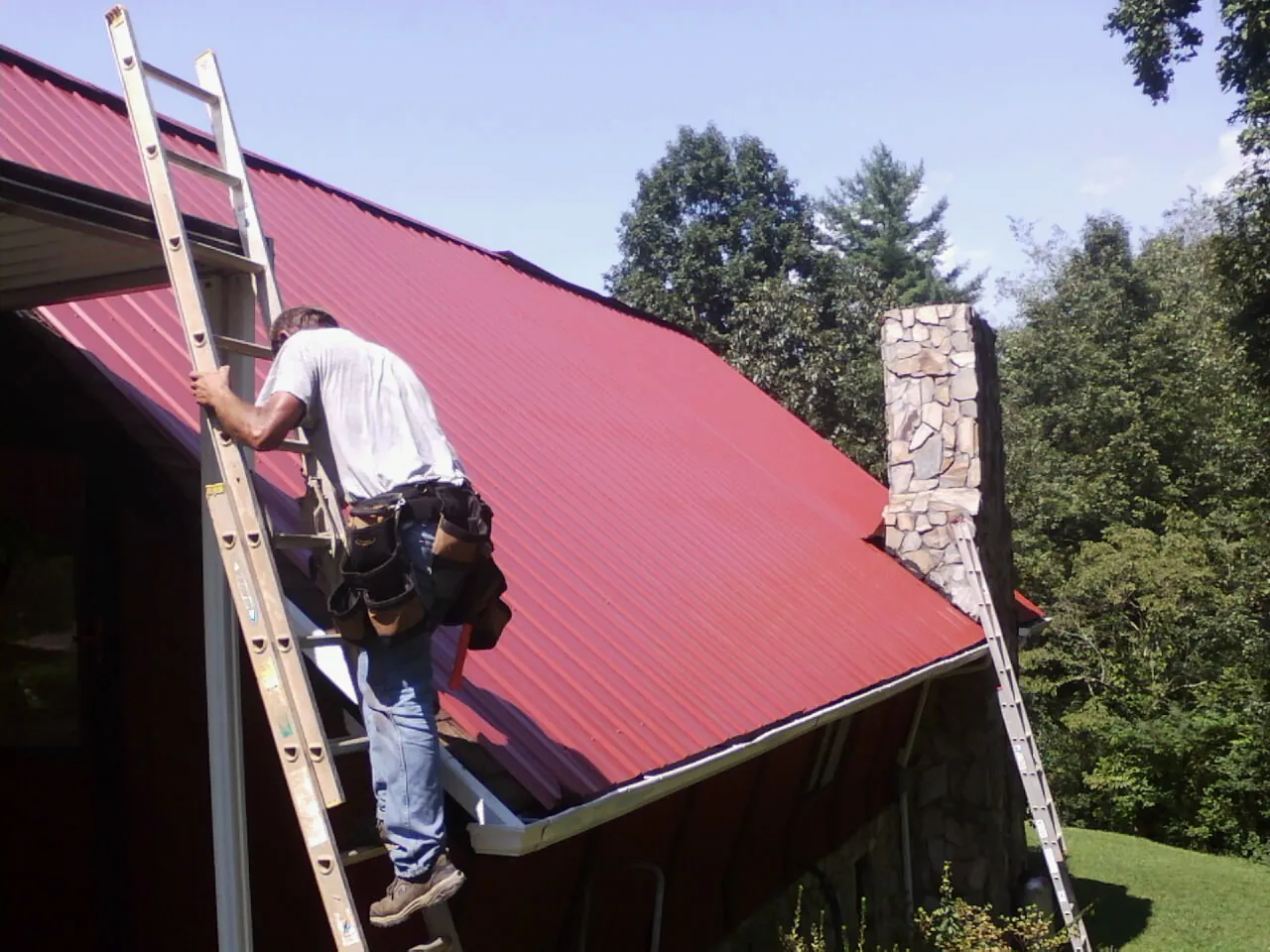 Roofer on a ladder beside a red metal roof and stone chimney — Soffit & Fascia Repair by Roofing Brothers in Cologne